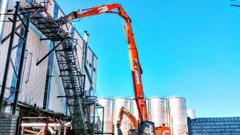 A high-reach demolition excavator fitted with a hydraulic shear, working immediately adjacent to an operational facility.